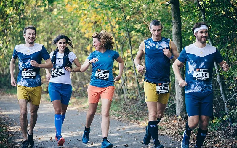 Vijf hardlopers met startnummers joggen samen over een bosrijk pad, glimlachend en pratend. Ze dragen sportkleding en het zonlicht schijnt door de bomen, wat een vrolijke sfeer creëert.